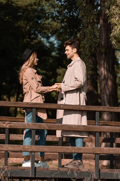 Profile of stylish couple in trench coats holding hands on wooden bridge — Stock Photo