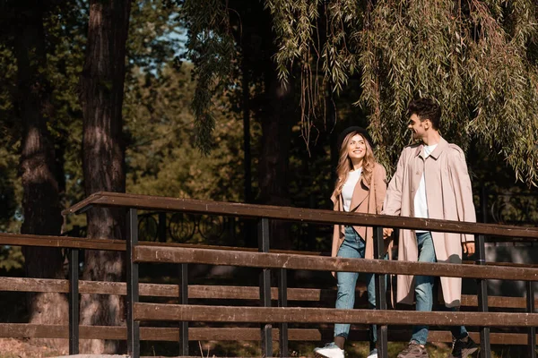 Man and woman in trench coats holding hands and walking on wooden bridge — Stock Photo