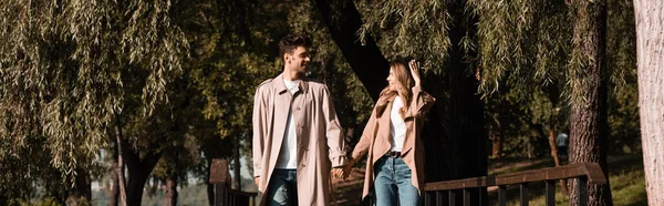 Panoramic concept of woman touching straw hat and looking at boyfriend in trench coat — Stock Photo