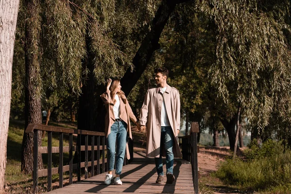 Woman touching straw hat and looking at boyfriend while walking on wooden bridge — Stock Photo