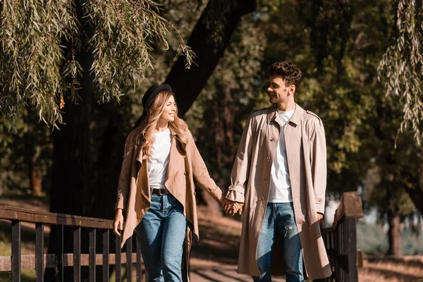 Couple in trench coats holding hands, looking at each other and walking on wooden bridge — Stock Photo