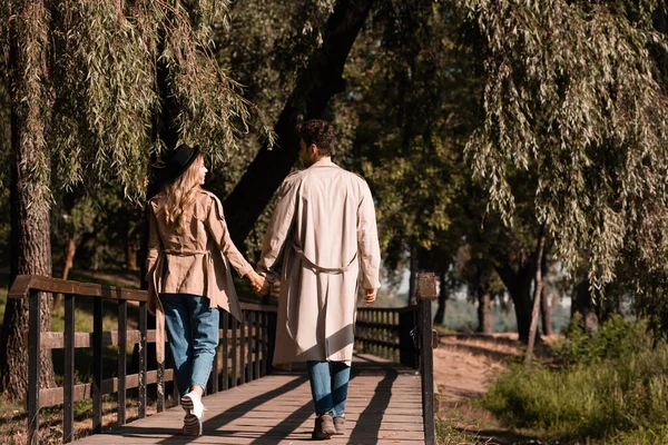 Casal em casacos de trincheira de mãos dadas e andando na ponte de madeira no parque outonal — Fotografia de Stock