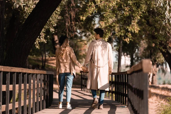 Man and woman in trench coats holding hands, looking at each other and walking on wooden bridge — Stock Photo