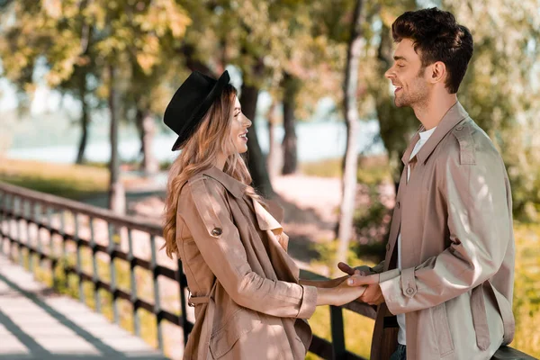 Vista lateral del hombre y la mujer en abrigos de trinchera tomados de la mano y mirándose mientras están de pie en el parque otoñal - foto de stock