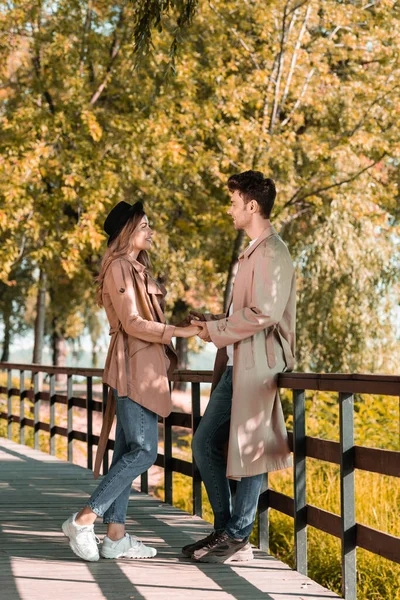 Профиль пары в плащах, держащихся за руки и смотрящих друг на друга, стоя в осеннем парке — стоковое фото