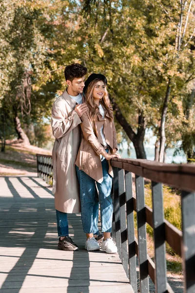 Man touching shoulders of woman in hat and trench coat standing on wooden bridge in autumnal park — Stock Photo