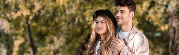 Panoramic crop of man touching shoulders of woman in hat and trench coat in autumnal park — Stock Photo