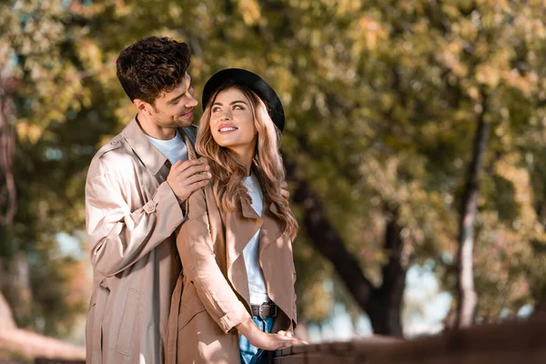 Selective focus of man touching shoulders of woman in hat and looking at each other in autumnal park — Stock Photo