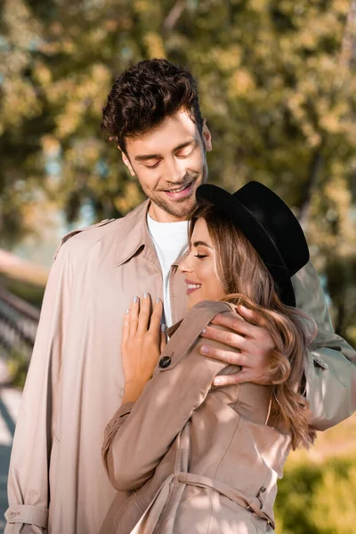 Hombre en gabardina abrazando a mujer en sombrero afuera - foto de stock