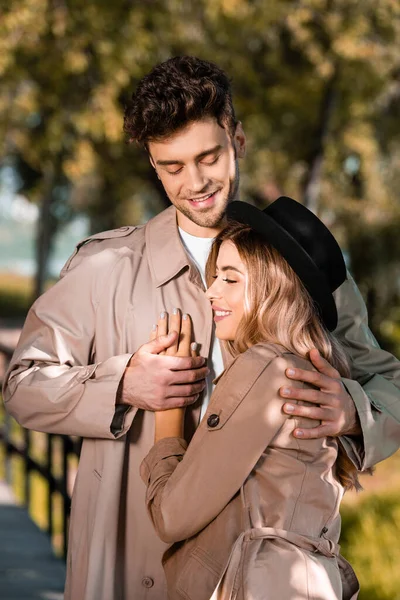 Hombre en gabardina abrazando y tocando la mano de mujer en sombrero exterior - foto de stock