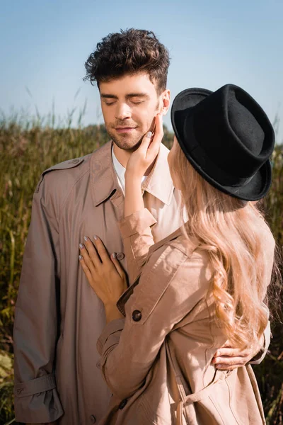 Femme blonde au chapeau touchant le visage de l'homme en trench coat à l'extérieur — Photo de stock