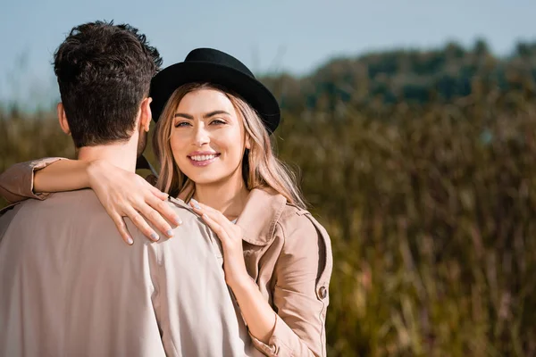 Blonde und fröhliche Frau mit schwarzem Hut umarmt Mann im Trenchcoat — Stockfoto