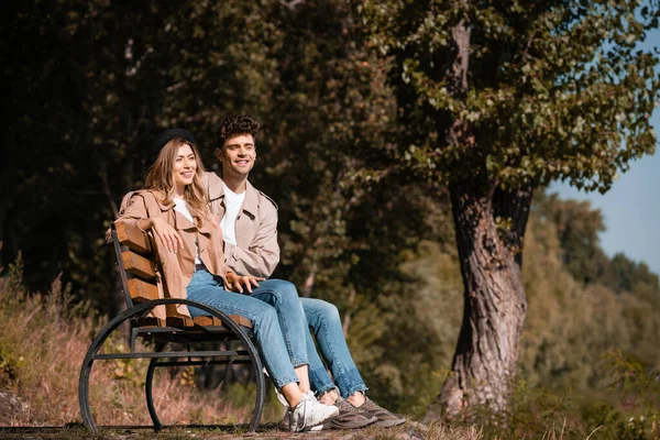 Femme blonde en chapeau et homme en trench coat assis sur le banc dans le parc — Photo de stock