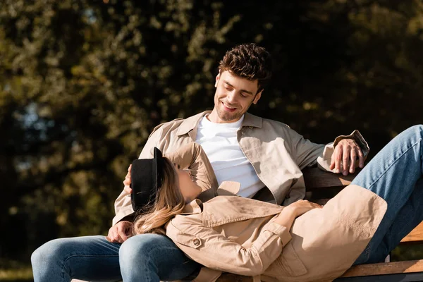 Mujer rubia en sombrero y hombre mirándose mientras descansan en el banco en el parque - foto de stock