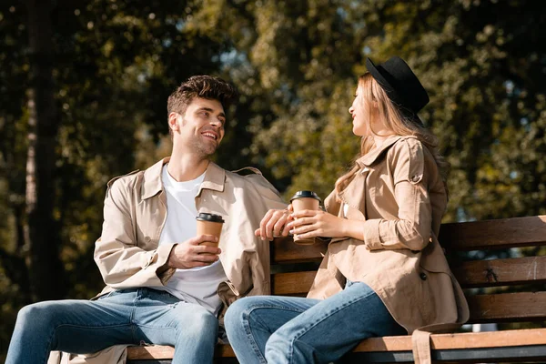Блондинка в шляпе и мужчина в плаще держат бумажные стаканчики, сидя на скамейке — стоковое фото