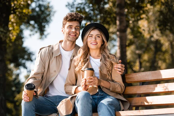 Femme en chapeau et homme en trench coat tenant des tasses en papier assis sur le banc — Photo de stock