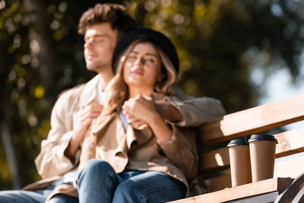 Selective focus of paper cups near blonde woman in hat and man in trench coat sitting on bench — Stock Photo