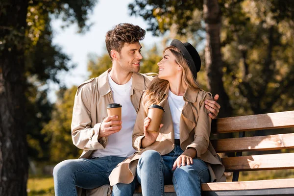 Blondine mit Hut und Mann im Trenchcoat halten Pappbecher in der Hand, während sie auf der Bank sitzen und in die Kamera schauen — Stockfoto