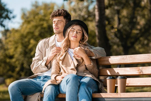 Femme blonde en chapeau et homme en trench coat assis sur un banc près de tasses en papier — Photo de stock