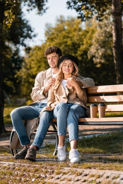 Femme blonde en chapeau et homme en trench coat assis sur un banc en bois près de tasses en papier — Photo de stock