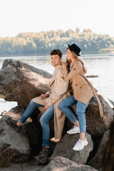 Mulher loira de chapéu olhando para o namorado e sentado em pedras perto do lago — Fotografia de Stock