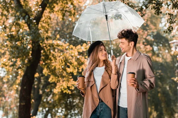 Casal em casacos de trincheira de pé sob guarda-chuva e segurando copos de papel com café para ir — Fotografia de Stock