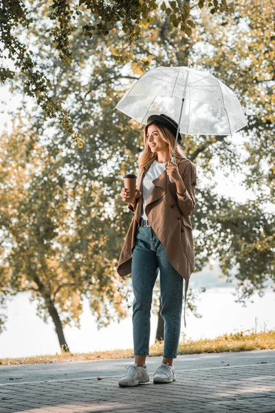 Woman in trench coat and hat standing under umbrella and holding paper cup with coffee to go in autumnal park — Stock Photo