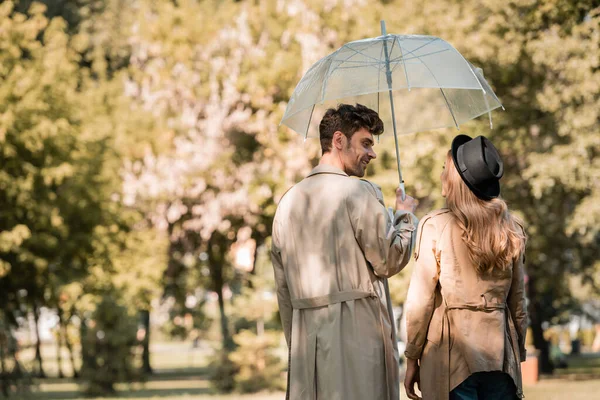 Woman in hat and man in trench coat standing under umbrella while looking at each other in autumnal park — Stock Photo