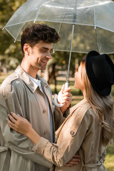 Man in trench coat standing under umbrella and looking at blonde woman in hat in autumnal park — Stock Photo