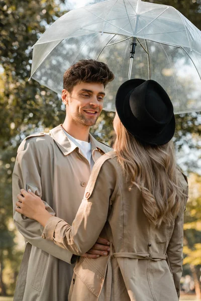 Mann im Trenchcoat steht unter Regenschirm und blickt Frau mit schwarzem Hut im herbstlichen Park an — Stockfoto