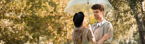 Cultivo panorámico de mujer en sombrero y hombre en gabardina de pie bajo paraguas en el parque otoñal - foto de stock