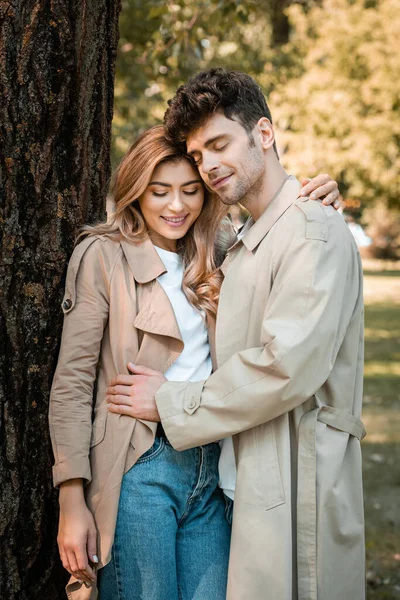 Pleased man with closed eyes hugging girlfriend in trench coat in autumnal park — Stock Photo