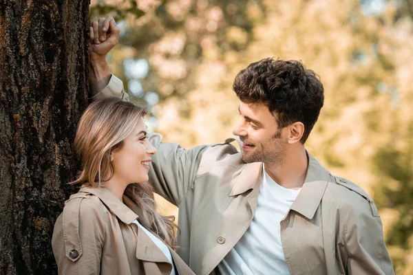 Boyfriend and girlfriend in trench coats looking at each other near tree trunk — Stock Photo