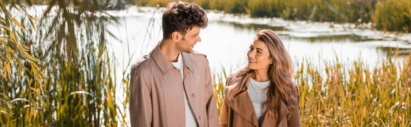 Prise de vue panoramique de la femme heureuse et l'homme en trench manteaux regardant les uns les autres près du lac dans le parc automnal — Photo de stock