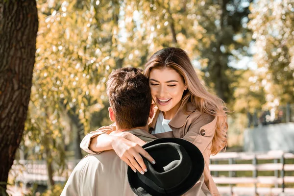 Mujer emocionada sosteniendo sombrero y abrazando novio en el parque otoñal - foto de stock
