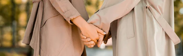 Panoramic crop of man and woman in trench coats holding hands in park — Stock Photo
