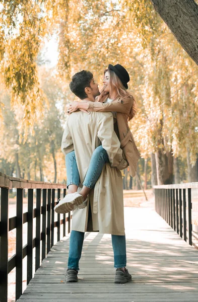 Man in trench coat holding in arms woman in hat while standing on wooden bridge in autumnal park — Stock Photo