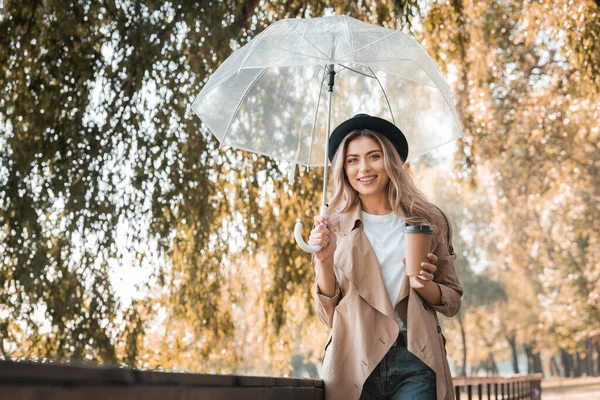 Блондинка в шляпе держит зонтик и бумажную чашку с кофе, чтобы пойти в осенний парк — стоковое фото
