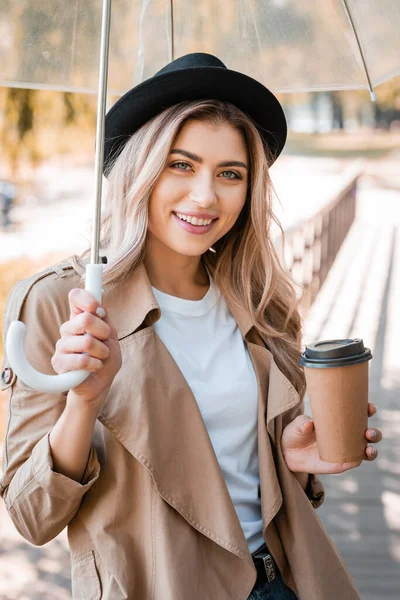 Femme en chapeau tenant parapluie et tasse en papier avec café pour aller dans le parc automnal — Photo de stock