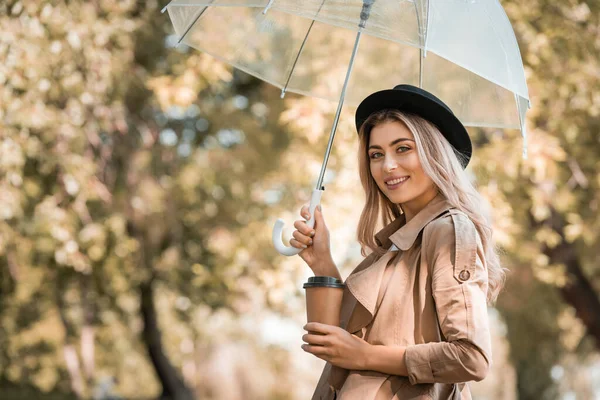 Mulher loira de chapéu segurando guarda-chuva e copo descartável com café para ir no parque outonal — Fotografia de Stock