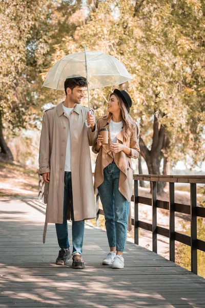 Couple in trench coats walking on wooden bridge under umbrella with coffee to go — Stock Photo