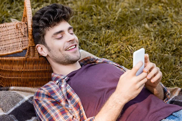 Mann schaut auf Smartphone, während er auf Decke in der Nähe von Weidenkorb liegt — Stockfoto