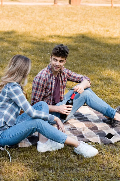 Мужчина держит бутылку красного вина рядом с женщиной, сидящей на клетчатом одеяле возле смартфона в парке — стоковое фото