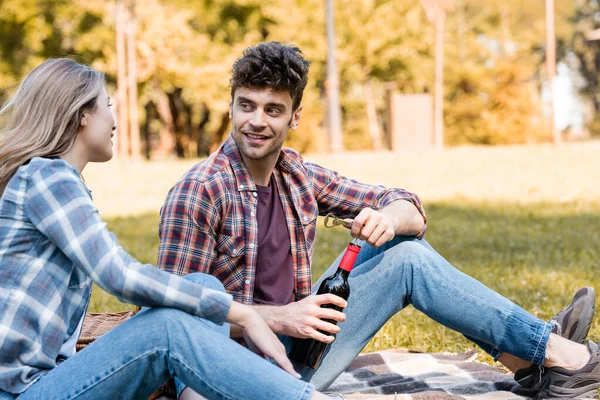 Мужчина держит бутылку красного вина рядом с женщиной, сидящей на клетчатом одеяле в парке — стоковое фото
