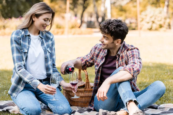 Homem segurando garrafa e derramando vinho tinto em vidro perto de mulher sentado em manta xadrez no parque — Fotografia de Stock