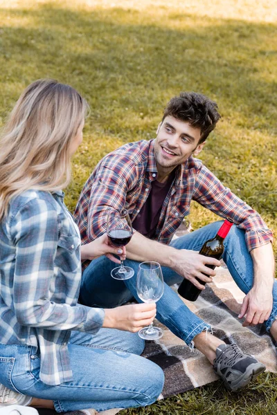Чоловік тримає пляшку червоного вина і дивиться на жінку в окулярах, сидячи на плетеній ковдрі в парку — стокове фото