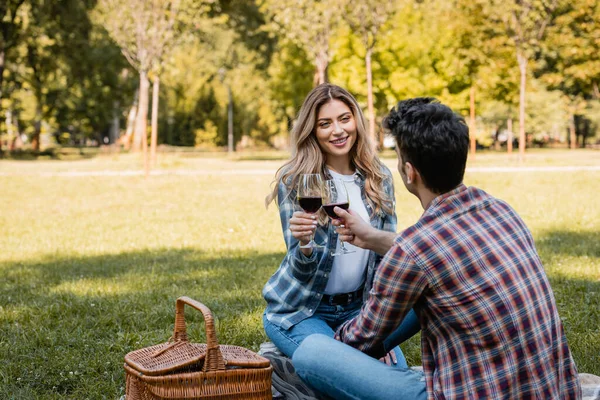 Man and woman sitting on blanket and holding glasses of red wine while clinking in park — Stock Photo