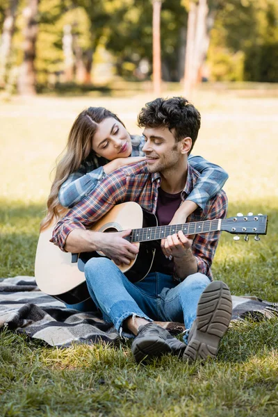 Mujer sentada en manta a cuadros y tocando novio tocando la guitarra acústica - foto de stock
