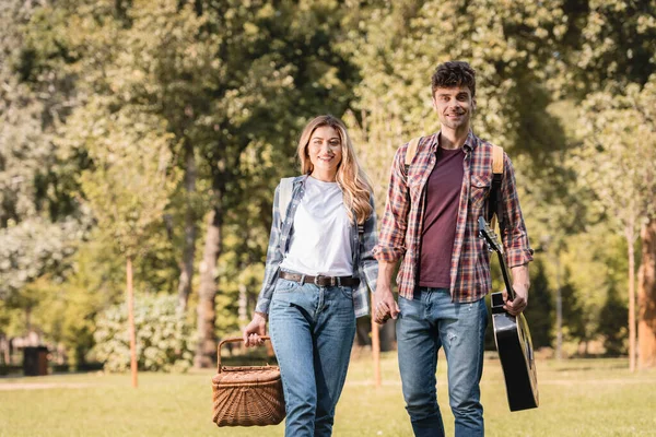 Boyfriend with acoustic guitar and girlfriend with wicker basket holding hands outside — Stock Photo
