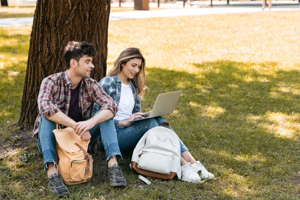 Hombre mirando a la computadora portátil mientras está sentado cerca de la mujer bajo el tronco del árbol - foto de stock